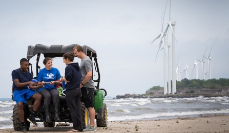 University at Buffalo students stand with a professor reviewing samples on a beach. Windmills are in the background. 