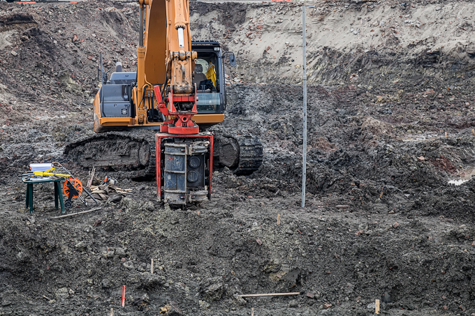 An industrial drill attached to construction vehicle similar to a digger is part-way in the ground. The ground is black and full of mud and clay. A table with additional equipment and gear sits off to the side. 