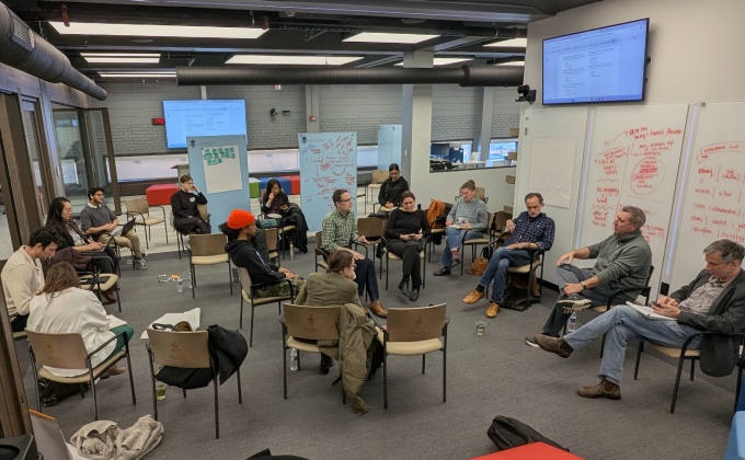 A group of about twenty people seated in a large circle of chairs in a modern conference or classroom space. Some people are speaking while others listen, take notes, or look down at papers. Whiteboards around the room are covered with handwritten notes and diagrams in red and black marker. Large wall‑mounted screens display documents. The room has exposed ceilings with bright rectangular light panels, glass walls on one side, and colorful cube-shaped seating in the foreground. Refreshments and trays of food are visible along the left side of the room. 