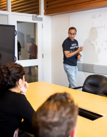A professor wearing a black "Avoid Negativity" T-shirt writes complex mathematical formulas on a whiteboard while speaking to a group of students seated around a conference table in a classroom or meeting room. A flat screen monitor is visible to the left, and reflections of people walking outside can be seen through a glass window. 