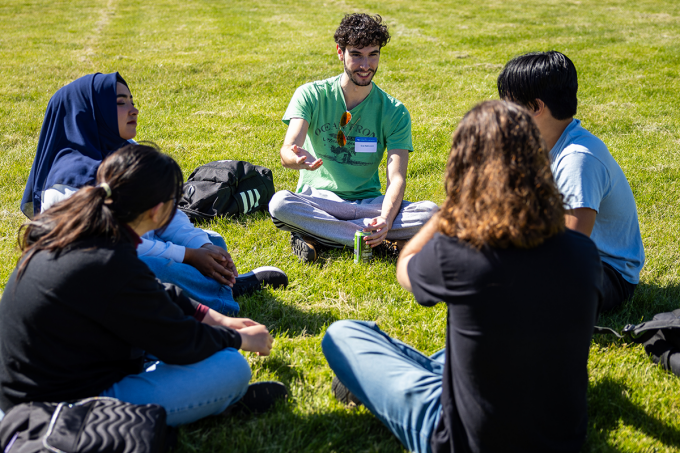 A small group of people sit in a circle on green grass outdoors, engaged in conversation. One person at the center gestures while speaking, holding a drink can, while others sit cross-legged or with legs extended, listening. Backpacks and personal items rest nearby, and the scene takes place in a sunny open lawn setting. 