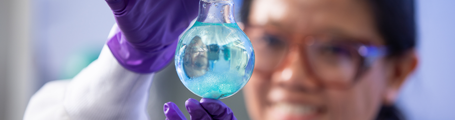 Student in full lab gear holds beaker in front of her face. 
