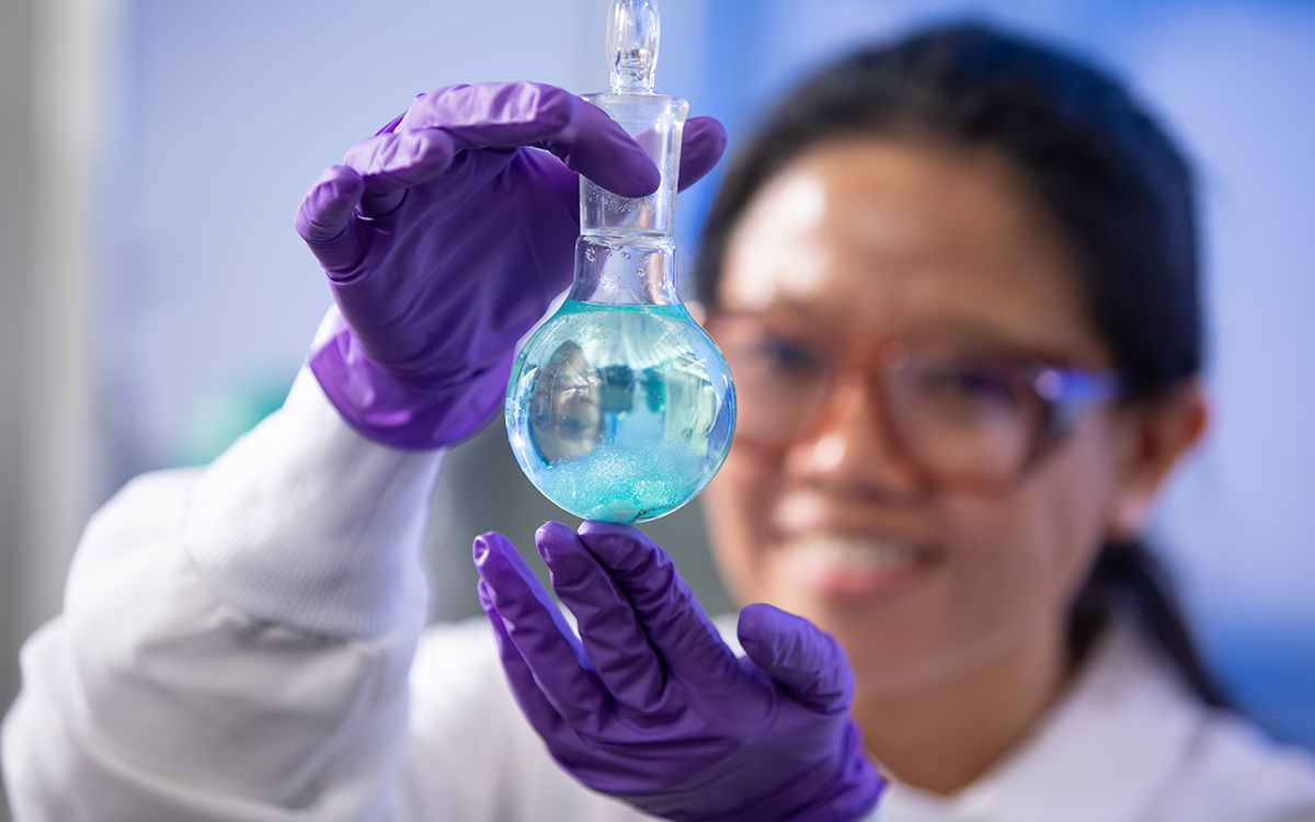 Student in full lab gear holds beaker in front of her face. 