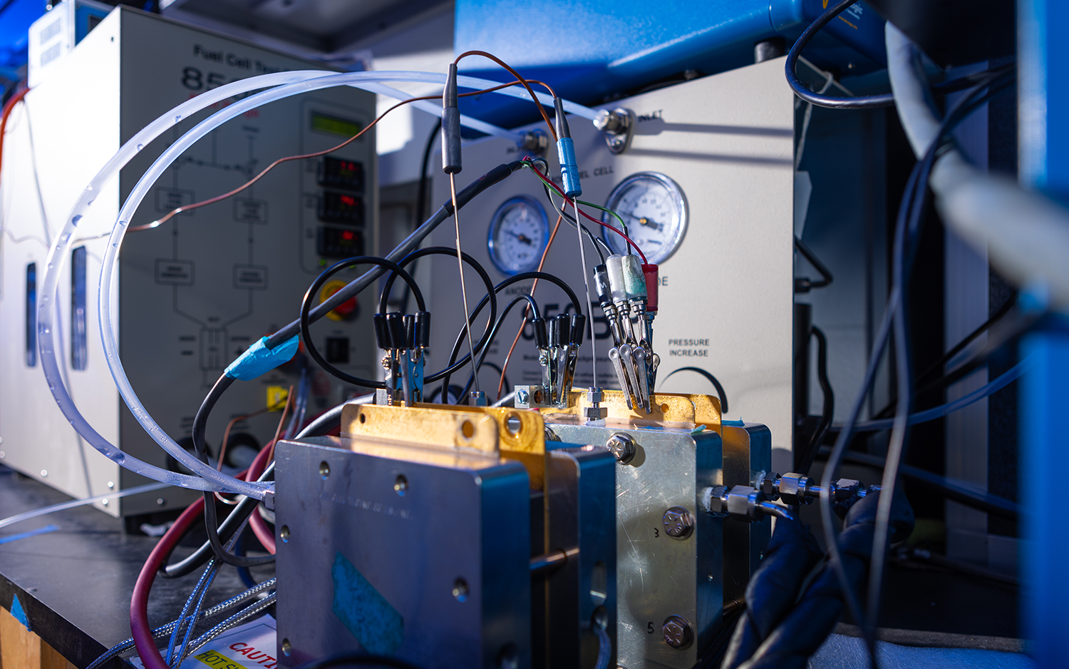 A device in a lab. Two boxes are connected to wires, tubes and different gauge readers. Behind them appear to be largeer boxes. 