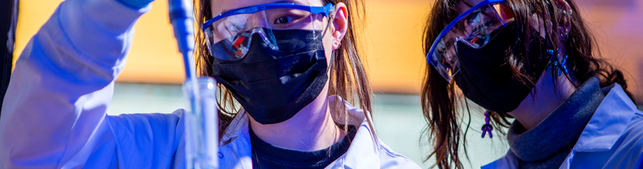 A student distributes wastewater from a needle into a test tube while a student observes behind the first student. Both students are dressed in lab coats, safety goggles, gloves and masks. 