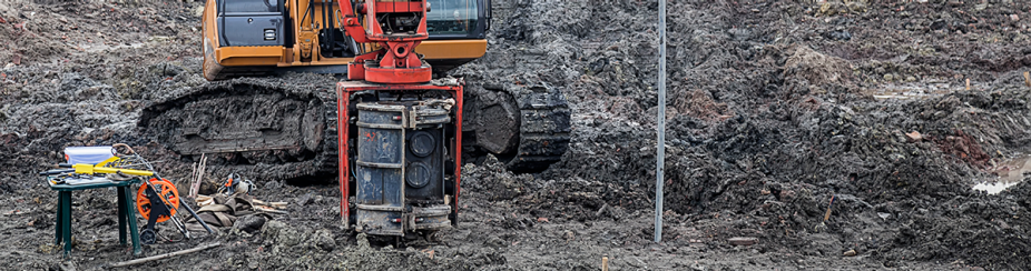 An industrial drill attached to construction vehicle similar to a digger is part-way in the ground. The ground is black and full of mud and clay. A table with additional equipment and gear sits off to the side. 