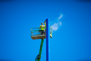 Two construction workers wearing safety gear and high-visibility clothing stand in a green aerial lift, working on a tall blue pole against a bright blue sky with a small cloud in the background. 