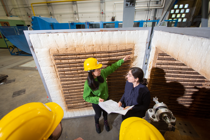 Two female researchers, a student wearing a hard hat and a faculty member, discuss experiments while a large open furnace sits behind them. There are two additional hardhats toward the bottom of the image.