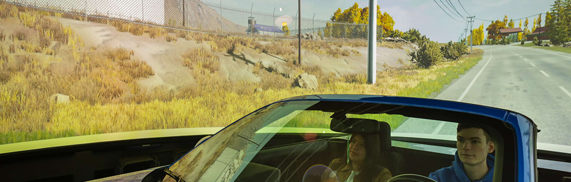 Two students sit in the front of a car; one male and one female. a ring of sceneic views wraps around them. It is the motion simulation laboratory. A spotlight shines from above. 
