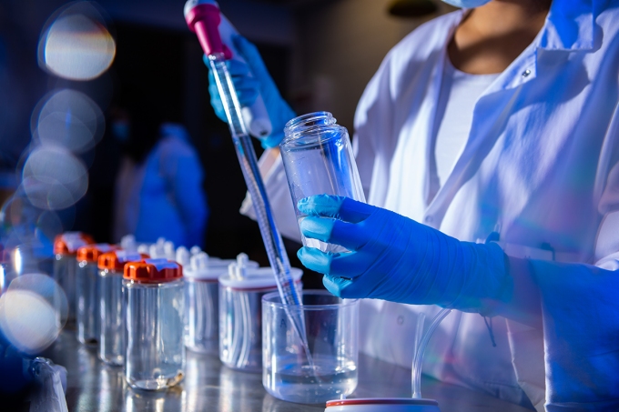 Tubes and water are in the front of the frame while a person wearing gloves and a lab coat drop samples into containers. 