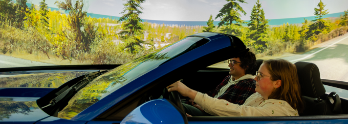 Two students sit in the front of a mustang in a driving simulator. The ring around them features a landscape of trees near a roadway. 