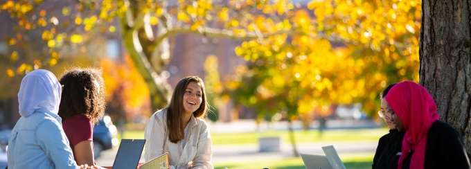 Students socialize in the grass near University at Buffalo's Ketter Hall. It is fall and the trees are starting to change colors. 