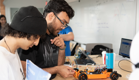 Two students stand at a table examining a small mechanical car that appears as if it could be autonomous. They are wearing t-shirts, and one has glasses and a beanie. Several other people are in the background. 