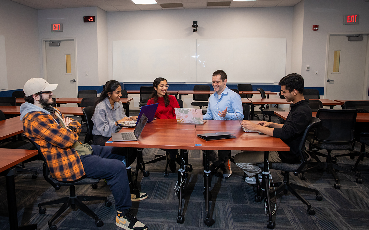 Four students and a faculty member sit at a table and laugh. 