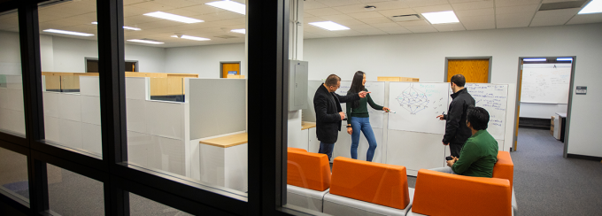 Three researchers stand in the foreground pointing and drawing on a whiteboard. A faculty member watches behind them drinking coffee. 