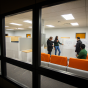 Three researchers stand in the foreground pointing and drawing on a whiteboard. A faculty member watches behind them drinking coffee. 