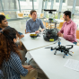 Four students and a faculty member sit around a table with several devices including a drone, some kind of tower and a headset on it. They are talking and smiling. Behind them, it appears to be a nice spring or summer day. 