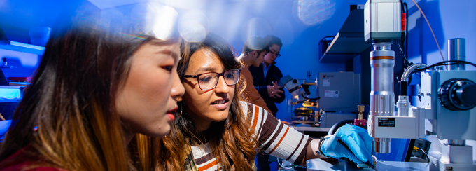 Two female students examine a specimen under a device that looks similar to a microscope. 