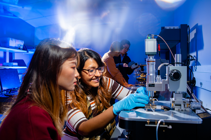 Two female students hold material near a tool that looks like an industrial microscope or a key maker. They are in the foreground while a male faculty member points to another device standing next to a male student. 