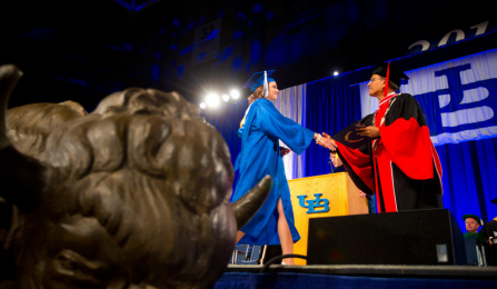 A student in a blue graduation cap and gown accepts a diploma as she crosses the stage.