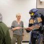 Professor exams a binder with a person dressed in full armor. It appears to be armor similar to what is worn by characters in the Halo video game. This is science-fiction space armore. Students stand in a classroom looking at the two in the front. 