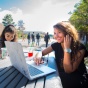 Two students sitting outside at table, one of whom is using a computer. 