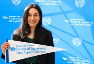 The first place winner Ghazal Vasseghi standing in front of the UB Photo wall holding a School of Engineering & Applied Sciences Banner and smiling. 