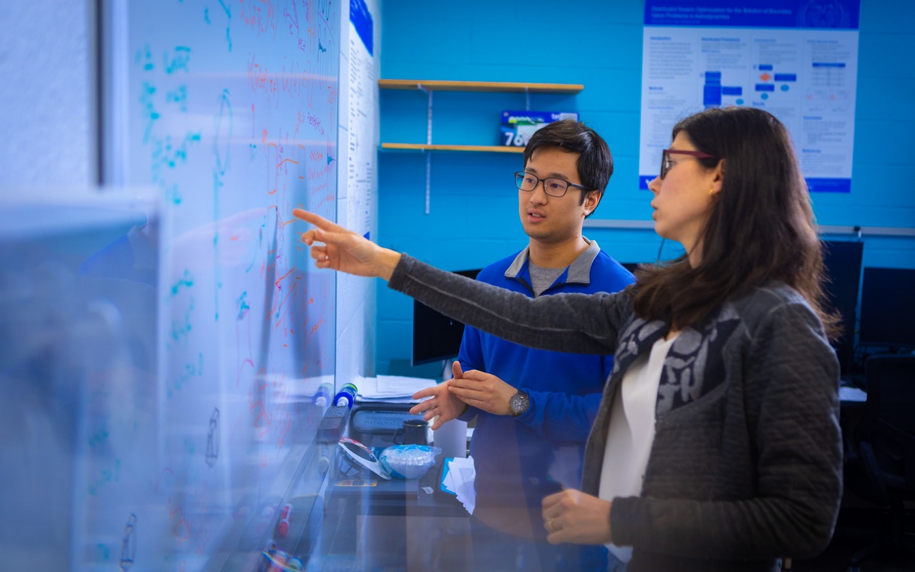 Two faculty members discuss something in front of a white board. 