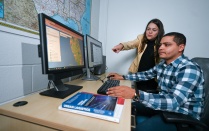 A student presenting as female discusses her research poster while holding a 3-D printed model of her research in one hand. 