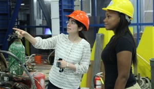 A student presenting as female discusses her research poster while holding a 3-D printed model of her research in one hand. 