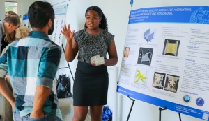 A student presenting as female discusses her research poster while holding a 3-D printed model of her research in one hand. 