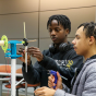 Two people work together in a classroom or lab space assembling a small mechanical device mounted on a vertical stand. The device includes a gear mechanism, rods, and wiring connected to a small motor or sensor. One person adjusts a component while the other holds a hot glue gun nearby. Tables, chairs, and an illuminated exit sign are visible in the background, indicating an instructional setting. 