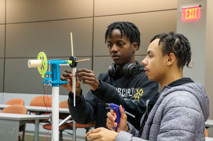 Two people work together in a classroom or lab space assembling a small mechanical device mounted on a vertical stand. The device includes a gear mechanism, rods, and wiring connected to a small motor or sensor. One person adjusts a component while the other holds a hot glue gun nearby. Tables, chairs, and an illuminated exit sign are visible in the background, indicating an instructional setting. 