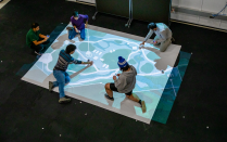 Four students and a faculty member sit on the ground. Below them is a rectangular sheet with a map projected on top of it. The students are pointing to different spots on the map while the faculty member holds a remote control off to the side. 