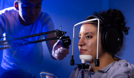A researcher adjusts equipment next to another researcher acting as a participant, wearing large headphones and seated with her chin and forehead positioned in a headrest. The setup is used for a precise audio or neurological study, lit with dramatic blue lighting. 