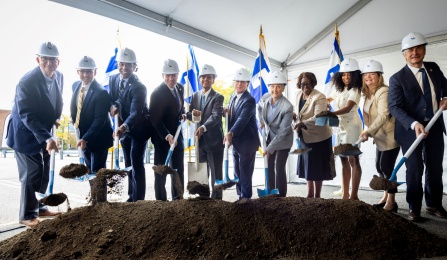 Group of people wearing hard hats and holding shovels in front of pile of dirt. 