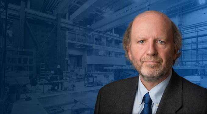 Michel Bruneau looks forward while the earthquake engineering lab sits behind him. He is overlayed on the image and two indidivuals are at a table reviewing a computer. Both wear hardhats, and one sits while the other stands. 