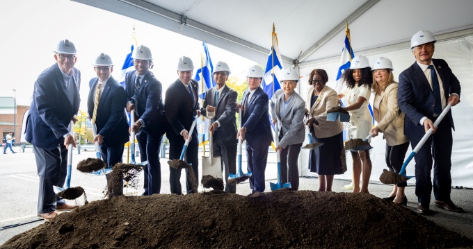 Group of people wearing hard hats and holding shovels in front of pile of dirt. 