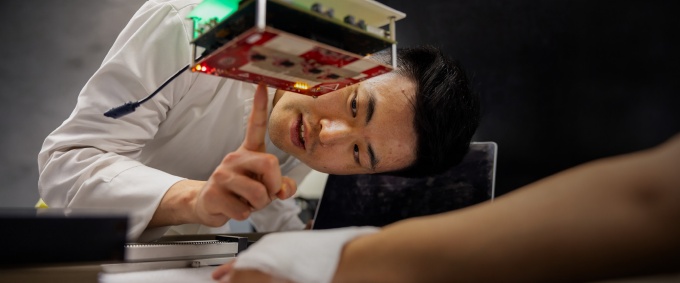 A person in a laboratory closely examines a prototype electronic device composed of stacked circuit boards mounted on metal standoffs. The person points toward illuminated components on the underside of the device while leaning over a workbench. A laptop and additional equipment are visible nearby, highlighting hands-on testing and development of electronics or embedded systems technology.