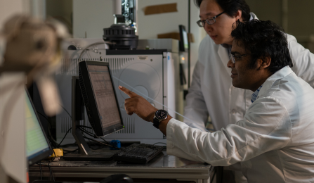 Miao Yu stands next to a student, both wear lab gear and point to a computer surrounded by equipment. 