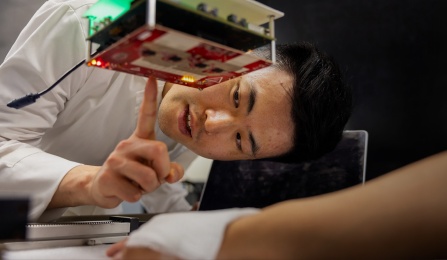 A person in a laboratory closely examines a prototype electronic device composed of stacked circuit boards mounted on metal standoffs. The person points toward illuminated components on the underside of the device while leaning over a workbench. A laptop and additional equipment are visible nearby, highlighting hands-on testing and development of electronics or embedded systems technology.