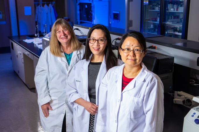 Photo of three women wearing white lab coats inside a research lab. 