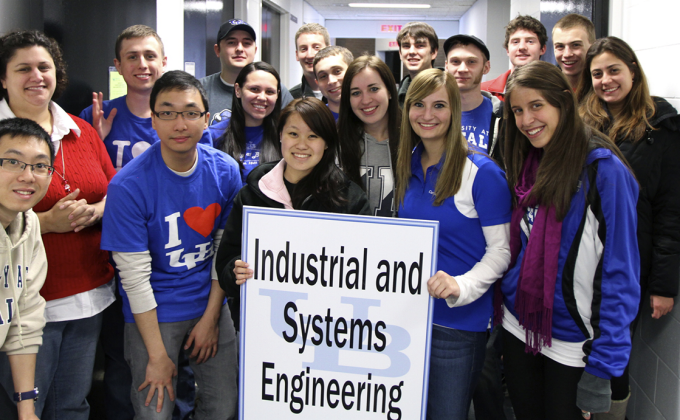 Sixteen students stand smiling while one hold a relative large sign featuring the UB logo (the letters "U" and "B" interconnected) with the Department name "Industrial and Systems Engineering" on top of the sign. 