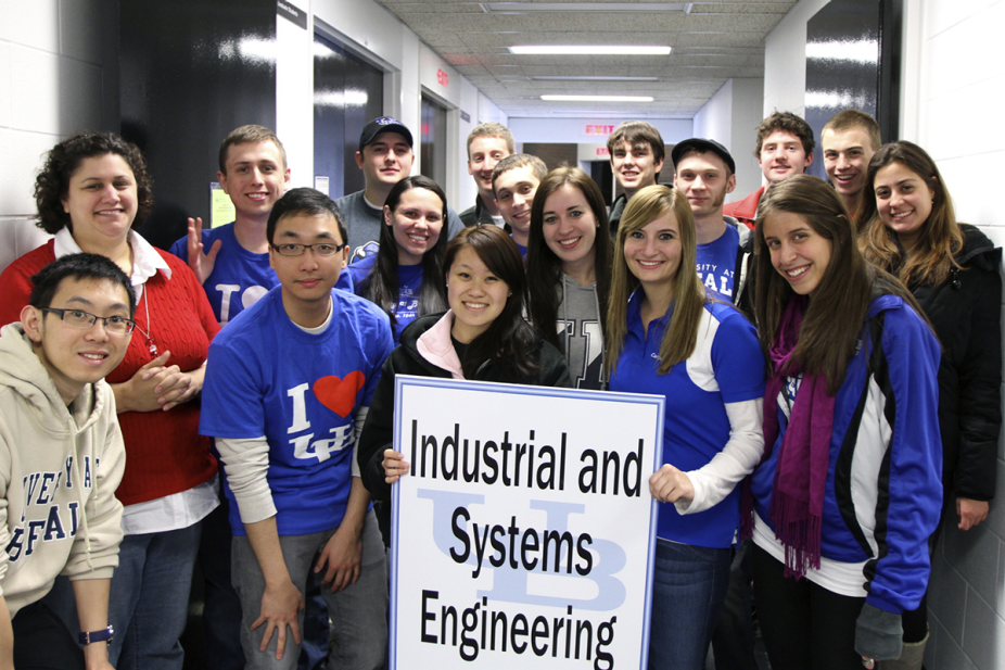 Sixteen students stand smiling while one hold a relative large sign featuring the UB logo (the letters "U" and "B" interconnected) with the Department name "Industrial and Systems Engineering" on top of the sign. 