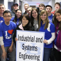 Sixteen students stand smiling while one hold a relative large sign featuring the UB logo (the letters "U" and "B" interconnected) with the Department name "Industrial and Systems Engineering" on top of the sign. 