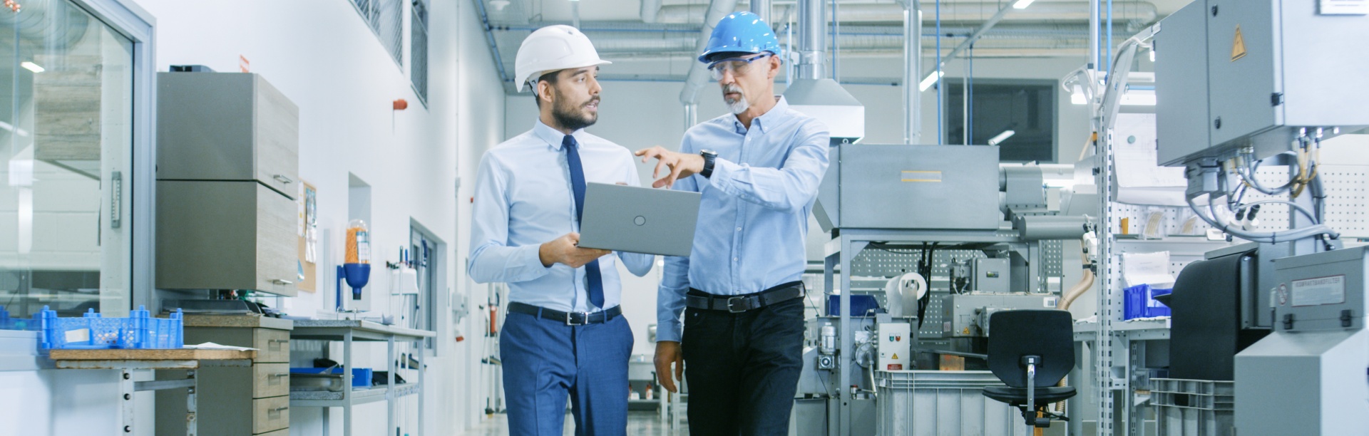 Two men in suits and hard hats holding laptop and having a discussion while walking through an industrial space. 