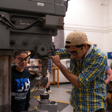 One student uses a drill press while another looks on. They appear to be drilling into a metal object. 