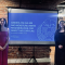 Four people stand on either side of a projection screen displaying a presentation titled &ldquo;Amberlite IRA-900 and Moisture Swing Regeneration for Direct Air Capture&rdquo; with &ldquo;University at Buffalo&rdquo; and &ldquo;Department of Chemical and Biological Engineering&rdquo; visible. The group is positioned indoors against an exposed brick wall with an arched opening, and the screen shows a blue slide with a buffalo emblem and the year &ldquo;1846.&rdquo;. 